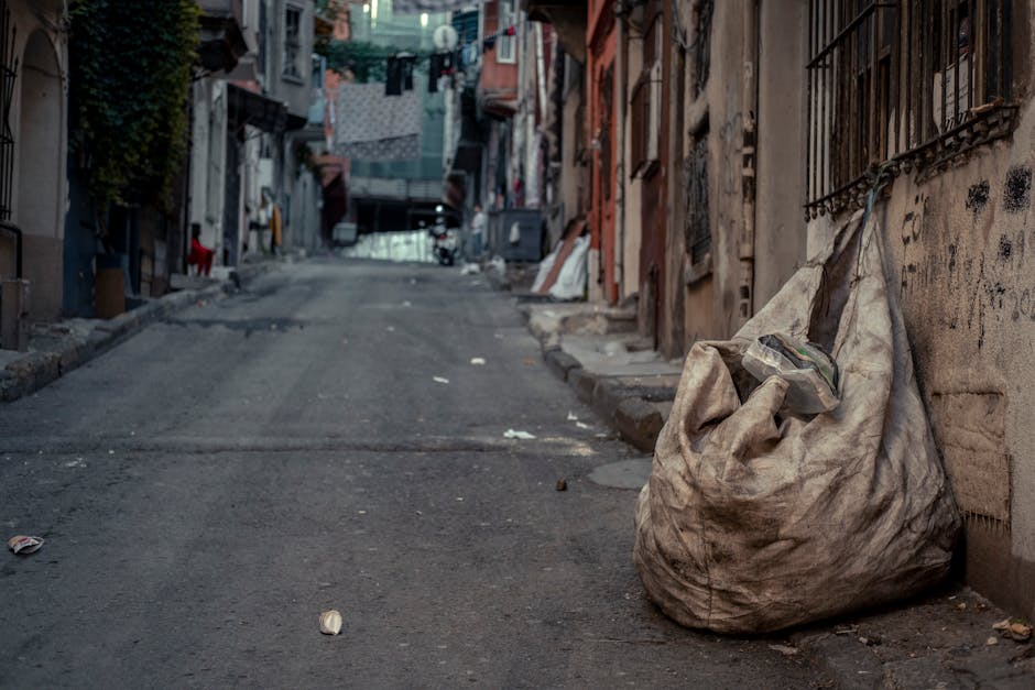 A large outdoor collection of mixed waste and rubbish overflowing from a grey metal recycling bin labeled for paper and cardboard, with various bags of black and coloured rubbish piled around it on a paved surface. The waste includes flattened cardboard boxes, crumpled paper, plastic bags, wrapping paper, and discarded packaging, some spilling onto the ground. To the left of the recycling bin, a small, silver hatchback car is parked behind a metal barrier, with a partially visible tree nearby. In the background, a commercial building with a blue scaffolding structure or construction fencing covers the upper floor, and storefronts with signage are visible beneath, including a fish bar. The scene appears to be in an urban parking area or street corner, illustrating a typical example of waste accumulation that private rubbish removal services, like those offered by Rubbish Removal Kensington, might assist with, especially as an alternative to council collection.