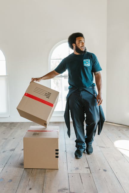 A man with dark skin, curly black hair, and a beard is standing in a bright, minimally decorated interior space with white walls, large arched window, and wooden flooring. He is wearing a navy blue T-shirt with a light blue patterned pocket, dark trousers, and has a jacket tied around his waist. The man is holding a medium-sized cardboard box in his right hand, which appears to be sealed with a red strip, and is placing it on top of a larger cardboard box positioned on the floor. The larger box is also sealed and has black tape on its edges. The scene suggests an indoor move or furniture removal process, with natural light illuminating the space through the window, emphasizing a clean, tidy environment suitable for private waste handling or rubbish removal services offered by Rubbish Removal Kensington.