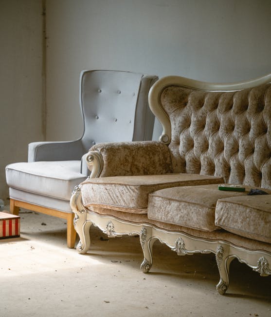 A close-up view of two vintage-style upholstered armchairs positioned side by side in an indoor space with a plain, slightly textured beige wall in the background. The chair on the left features a high, curved backrest with button-tufted detailing, covered in light grey or off-white fabric with a smooth, matte finish, and has wooden legs visible beneath the seat. The chair on the right has a rolled armrest, a high back with tufted upholstery in a beige or light brown fabric, and intricate carved wooden details along its frame and legs, which are painted in a soft, muted finish. Partially visible on the right side of the image is a small dark object resting on the armrest, possibly a remote control. The scene appears to be in a space used for furniture storage or a staging area, with a concrete or hardwood floor, and minimal natural or artificial lighting casting soft shadows. This setup hints at an environment where furniture may be temporarily stored or prepared for removal, aligning with the services of independent waste disposal or furniture clearance providers like Rubbish Removal Kensington.