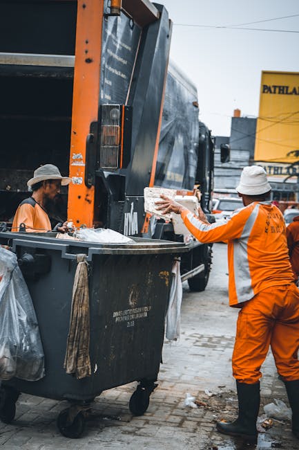 Two waste collection workers dressed in high-visibility orange uniforms and white hard hats are engaged in an outdoor rubbish removal operation. One worker is seen from behind, tipping a white, rectangular item into the large rear compartment of a black and orange waste collection vehicle. The other worker, standing on the left side, appears to be supervising or assisting, with a focus on the loading process. The waste truck features a large, enclosed, black metal body with reflective lights and safety markings, positioned on a paved area, possibly a driveway or street. In the background, there is a yellow building with black signage, and the scene is set in natural daylight, with an overcast sky evident. The setting suggests a practical, on-site disposal or clearance activity, consistent with alternatives to public rubbish collection, perhaps managed privately by companies like Rubbish Removal Kensington, serving residential or commercial clients seeking detailed, professional waste management services.