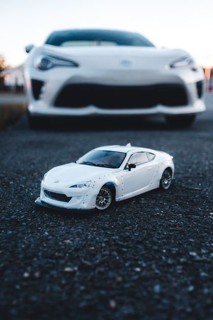 A close-up photograph of a small white remote-controlled or model sports car with a sleek, aerodynamic design, detailed with black-tinted windows, silver wheels, and a smooth, glossy finish. The car is positioned on a textured black asphalt or gravel surface, with the front of the car slightly angled toward the camera. In the background, a slightly out-of-focus larger white vehicle, likely an actual car, is parked on the same surface, with its lower front end visible and a blurred environment behind it. The lighting appears natural, with a soft, dusk-like atmosphere, casting gentle shadows on the ground and highlighting the contours of both vehicles. The scene suggests a setting where detailed car models or RC vehicles are showcased, and it subtly aligns with themes of specific vehicle handling or private car collection, in contrast to traditional rubbish removal services. Rubbish Removal Kensington would focus on the context of removing or handling waste related to automotive or garage decluttering, though the primary objects here are cars, not waste materials.