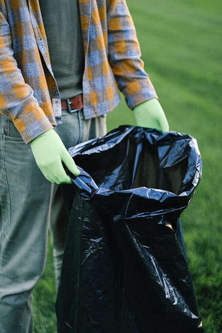 A person stands outdoors on a grassy surface, wearing a yellow and grey checkered shirt over a grey t-shirt, with green gloves on their hands. They are holding open a large black rubbish bag made of sturdy plastic, prepared for waste collection or removal. The background features a well-maintained lawn, indicating an outdoor setting suitable for rubbish disposal or garden waste collection. The scene portrays a professional waste management activity, consistent with private rubbish removal services such as those provided by Rubbish Removal Kensington. The natural lighting suggests daytime, and the focus on the open bag reflects the process of clearing or collecting rubbish in a controlled, environmentally conscious manner, aligning with the practice of independent collection or on-site clearance in residential or commercial settings.