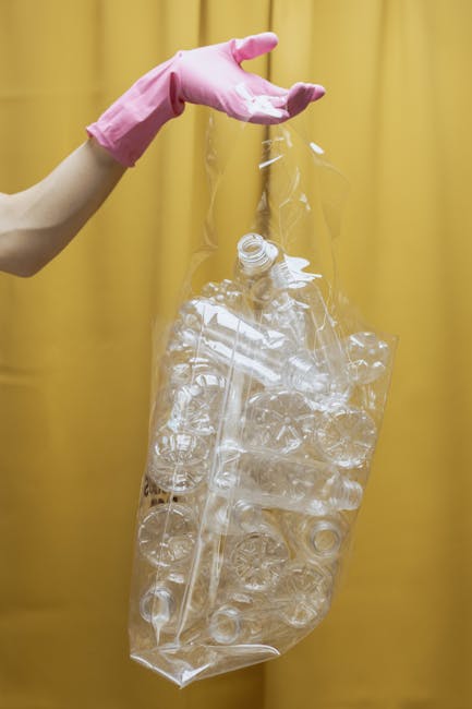 A person's arm wearing a pink rubber glove is holding a transparent plastic bag filled with crushed and intact clear plastic bottles and containers, set against a plain yellow backdrop. The bag appears to be lightweight and recyclable, with the bottles reflecting light and showing various shapes and sizes typical of plastic packaging waste. The scene emphasizes the process of collection or disposal of plastic rubbish, reflecting concepts of waste segregation or private waste handling outside of standard municipal rubbish collection. The focus on the plastic bottles and the neat arrangement within the bag highlights the importance of proper recycling and waste separation, which is relevant to independent rubbish removal services such as those offered by Rubbish Removal Kensington. The image's clean background and straightforward composition support a professional approach to waste management, underscoring the significance of environmentally responsible disposal practices with an emphasis on plastics, an important category within general rubbish removal activities.