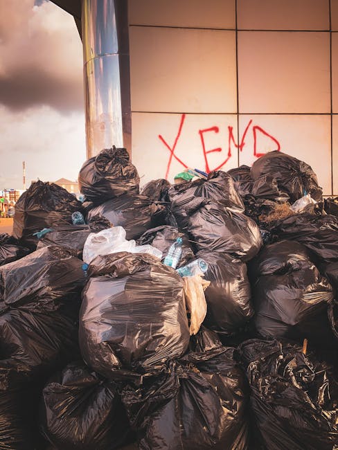 A large pile of black garbage bags filled with waste, stacked against a modern tiled wall on an urban pavement. Several clear plastic water bottles, some with labels and others without, are visible protruding from the bags. The bags appear tightly knotted at the top and are arranged haphazardly, some leaning against each other. Behind the waste, the wall features graffiti with the word 'XEND' spray-painted in red. The environment suggests an outdoor area designated for rubbish collection or disposal, possibly an alternative private waste removal site managed by services like Rubbish Removal Kensington, accommodating excess waste outside standard municipal collection. The lighting indicates late afternoon or early evening, with a cloudy sky partially visible in the background, enhancing the urban context suited for independent waste clearance activities. Overall, the scene illustrates a typical instance of private rubbish removal needs in a city setting, emphasizing the importance of professional waste management services around Kensington.