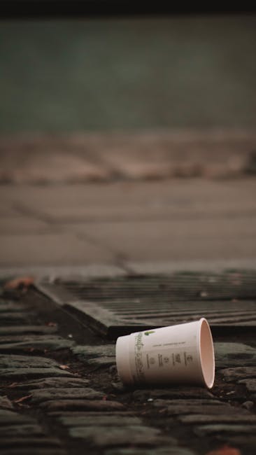 A discarded white paper cup with printed text lies on a weathered brick pavement, partially tilted and resting near a metal grate embedded into the ground. The pavement appears aged, with some gravel and small debris visible around the cup, suggesting a public outdoor area. In the background, a blurred greenish wall or fence is partially visible, indicating an exterior setting such as a street or courtyard. The scene is lit with natural, diffuse light, typical of an overcast day, which highlights the textures of the bricks and the cup. This image illustrates the type of waste that might be collected or removed during private or independent rubbish clearance services, such as those offered by Rubbish Removal Kensington, particularly focusing on small-scale litter or household waste disposal in urban outdoor environments.