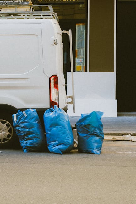 The image shows the rear side of a white commercial van parked on a paved street in an urban environment, with two large blue bulk bags filled with waste leaning against the vehicle's rear bumper. The bags are made of durable plastic with a slightly crinkled texture and are positioned in a row, one slightly to the left and the other in the middle, partially obscuring the van’s tail lights. The van is clean with a smooth finish, and a ladder rack is mounted on top, extending slightly beyond the roof. Behind the van, there is a storefront with a large window and a white step at the entrance, possibly indicating a commercial premises or waste collection point. The scene is lit by natural daylight, suggesting daytime, with the overall setting implying an on-site waste collection or rubbish removal activity managed by Rubbish Removal Kensington, emphasizing private waste handling outside of municipal disposal channels.