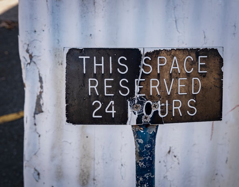 A weathered rectangular metal sign mounted on a rusted pole positioned against a white painted wall. The sign features white uppercase lettering that reads, 'THIS SPACE RESERVED 24 HOURS,' with some parts of the paint and surface chipped and corroded, revealing a dark underlying metal. The background includes the rough, slightly stained surface of the wall, and part of a faded yellow line is visible on the dark asphalt pavement to the left. The sign indicates a designated parking or loading zone, which may relate indirectly to private waste handling or on-site clearance activities conducted by rubbish removal services in Kensington, such as those provided by Rubbish Removal Kensington, ensuring efficient waste management outside traditional council collection hours. The overall scene emphasizes the utilitarian nature of urban signage associated with managing vehicle or service spaces for waste disposal or clearance, highlighting a typical element found near residential or commercial premises involved in rubbish removal and private disposal arrangements.